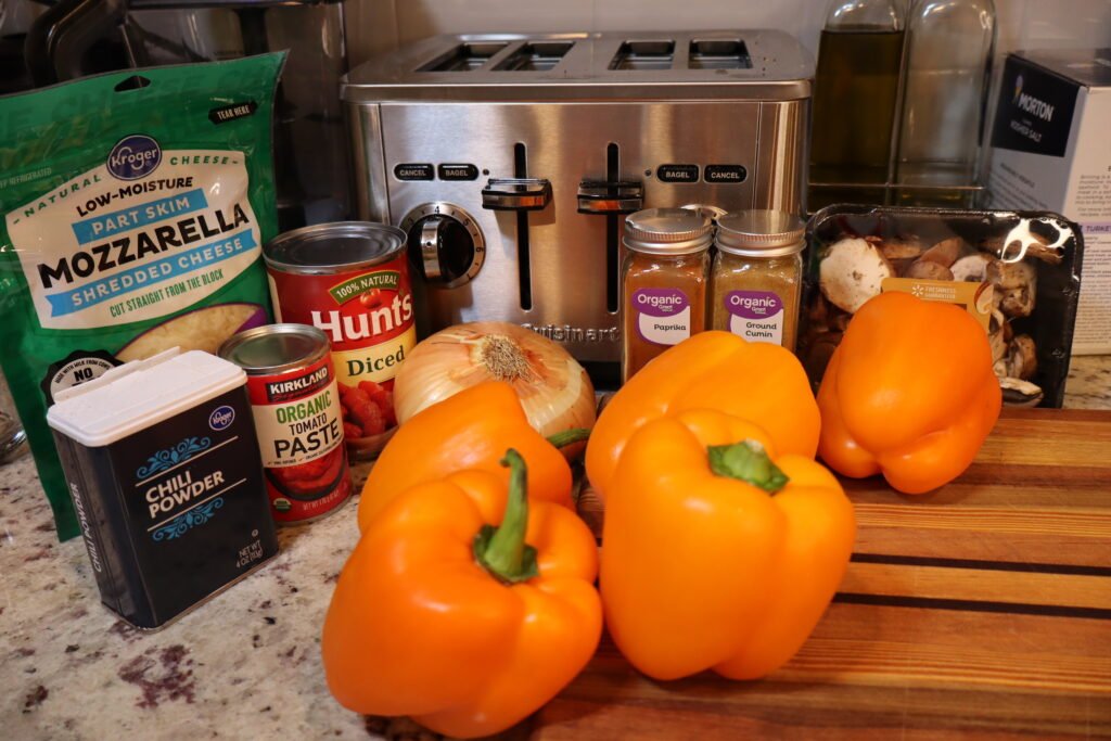 Ingredients on a counter that include bell peppers, baby bella mushrooms, shredded mozzarella, tomato paste, canned diced tomatoes, onion, and various spices.