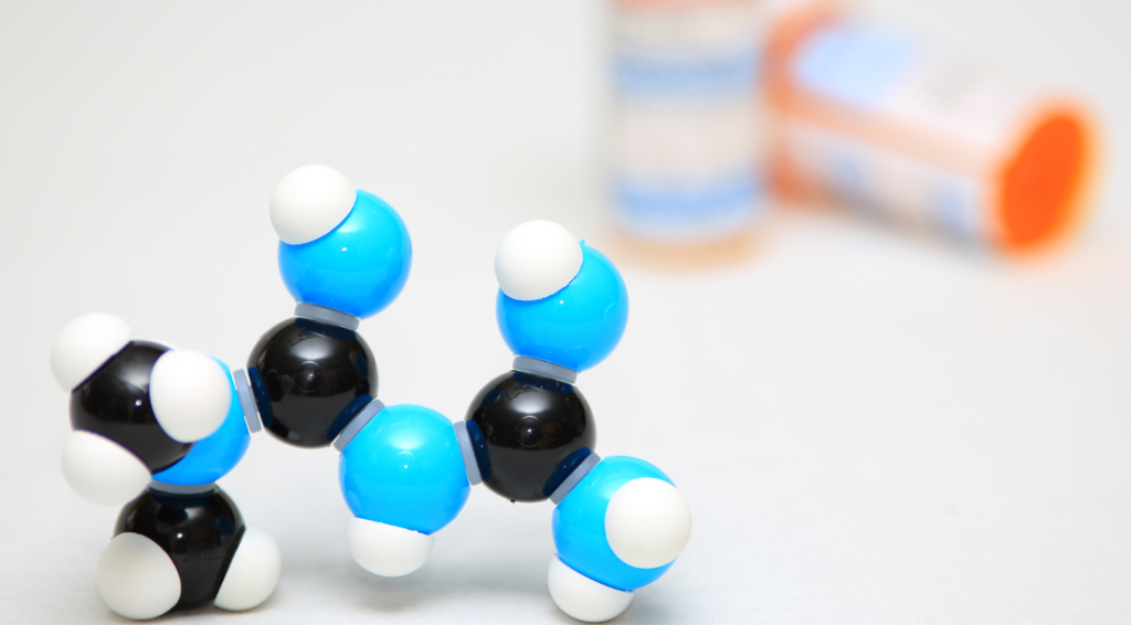 A chemical compound made of black, blue, and white balls is up close in focus, meant to represent Metformin. Behind it are two out of focus pill bottles.